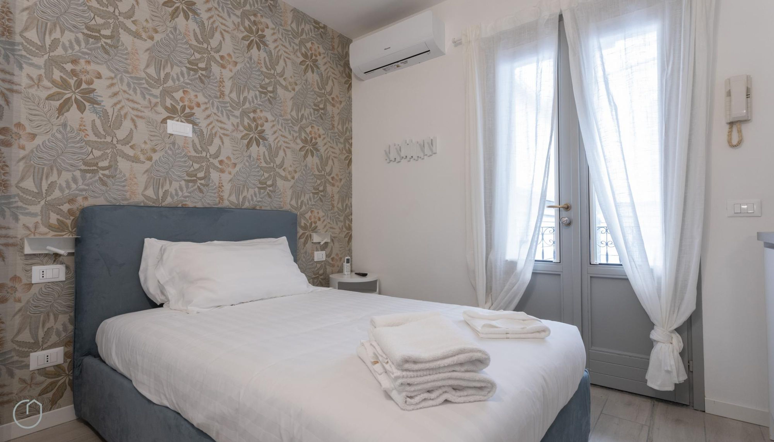 A neatly made bed with white linens and towels, a blue headboard, and a floral-patterned wallpaper.