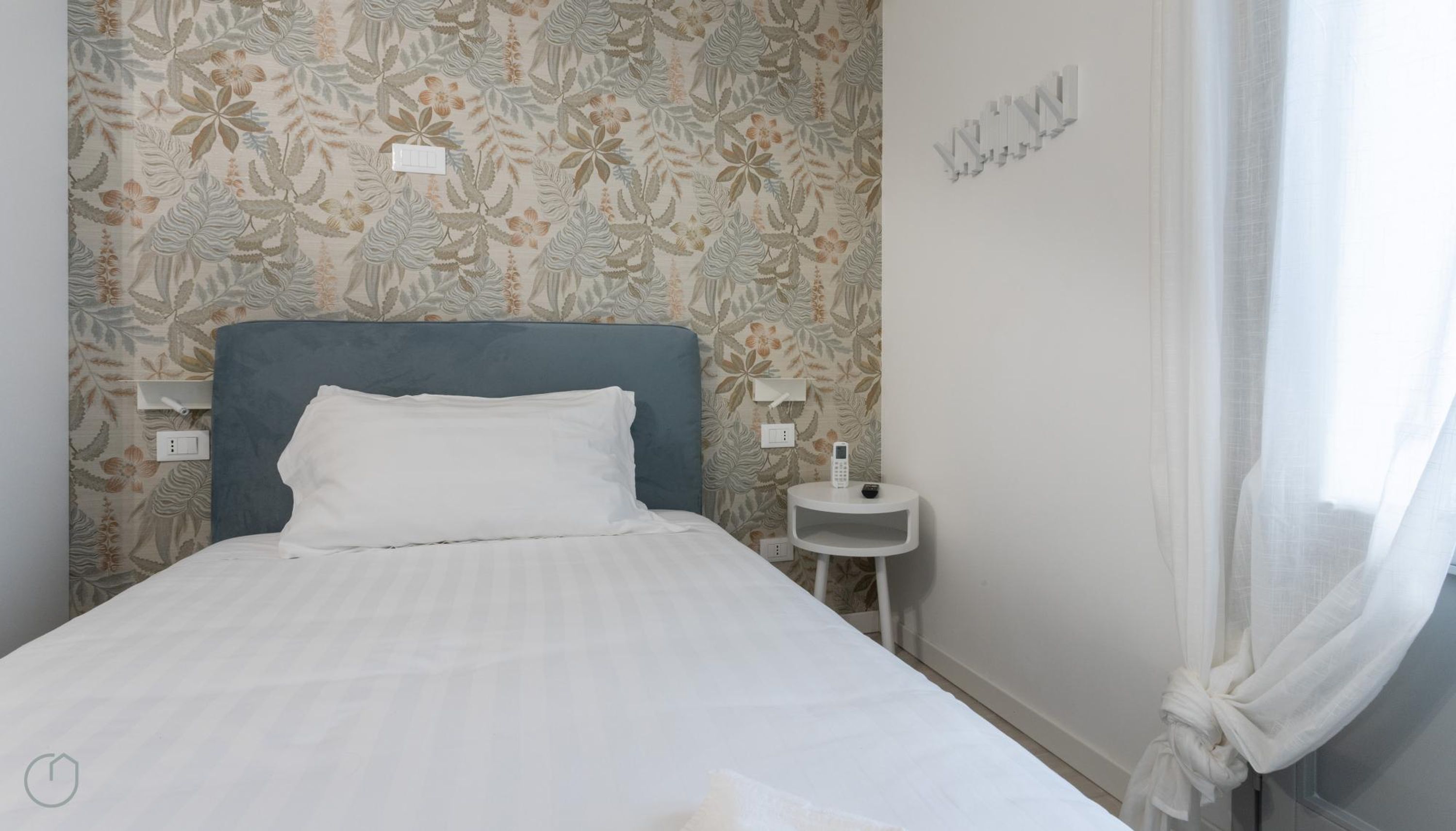 Cama bem arrumada com lençóis brancos, cabeceira azul e papel de parede com padrão floral. Uma pequena mesa de cabeceira com um telefone e um abajur são visíveis.