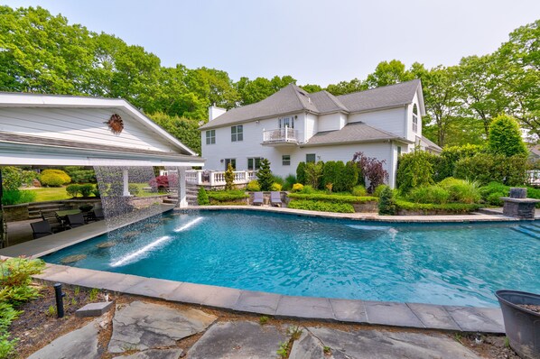 A large swimming pool with a stone deck, surrounded by a well-maintained garden and a two-story house with a balcony.