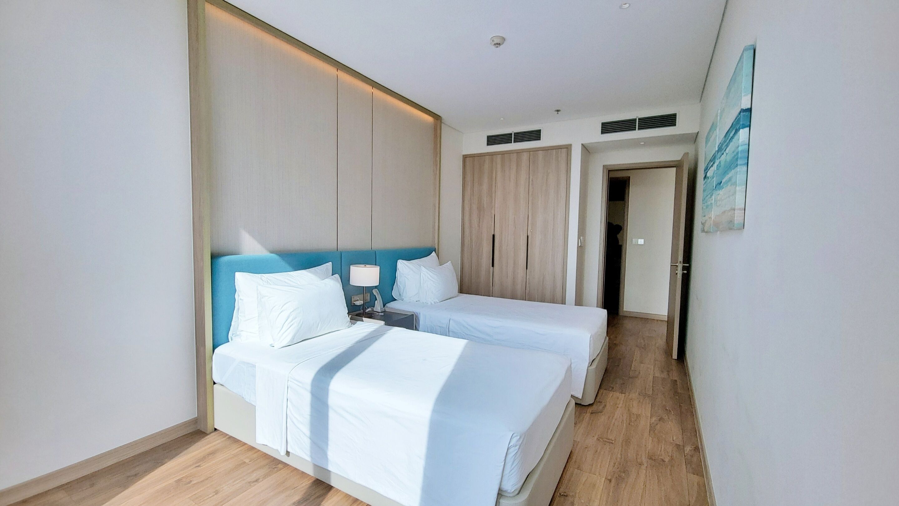 A hotel room with two beds, a blue headboard, a bedside table with a lamp, and a wardrobe.