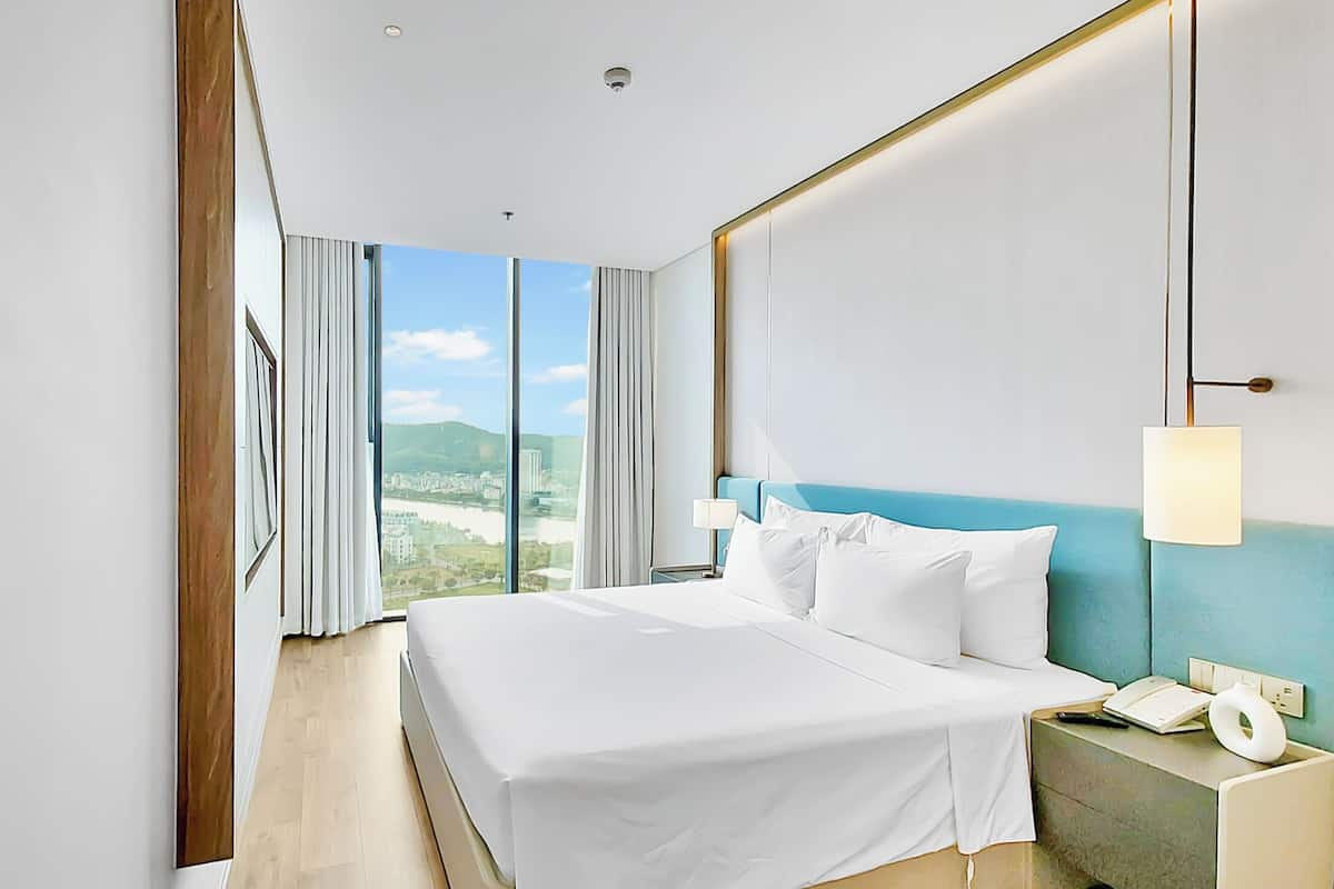 A hotel room with a large bed, a nightstand with a phone, and a view of a cityscape through large windows.