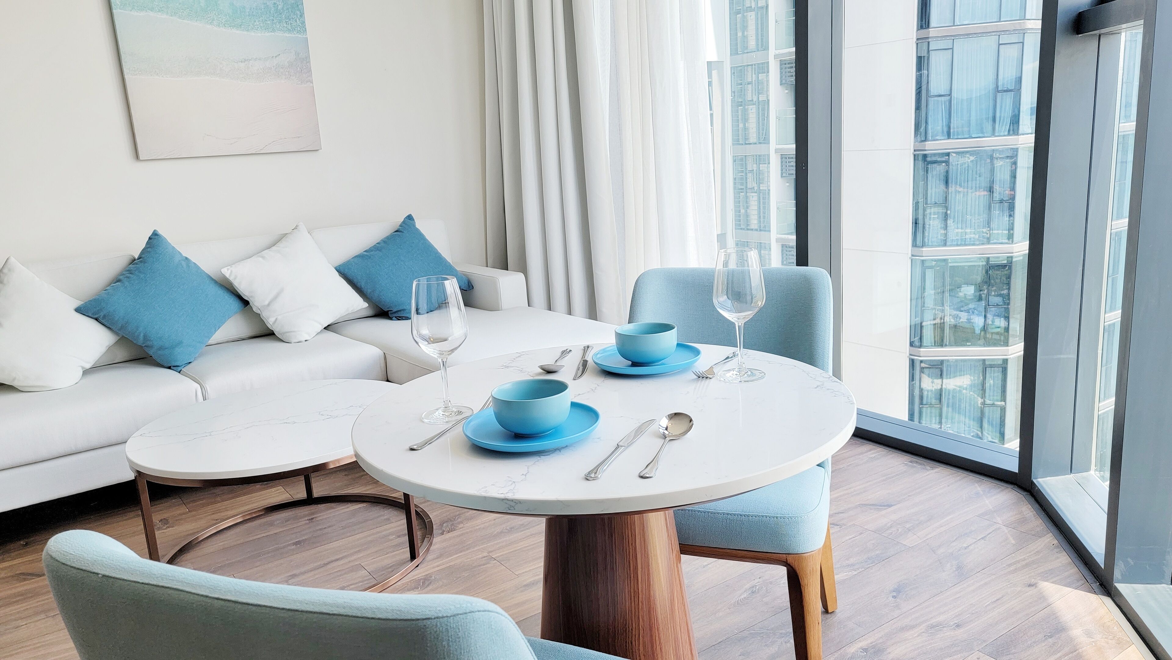 A modern living room with a white sofa, blue cushions, a round dining table with blue plates and silverware, and a view of a cityscape through large windows.