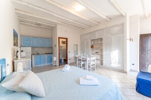 Quarto com cama, almofadas, mesa de cabeceira, escrivaninha e uma cadeira. Banheiro visível por uma porta aberta.