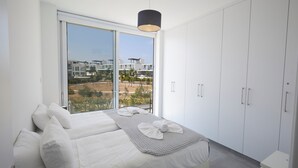 A modern bedroom with a large bed, white wardrobe, and a view of buildings through a sliding door.