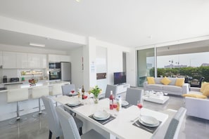 A modern kitchen with white cabinets, a dining area with a white table set for four, and a sofa with yellow cushions. - Protaras Olivine Villa Ol01 (Paralimni)