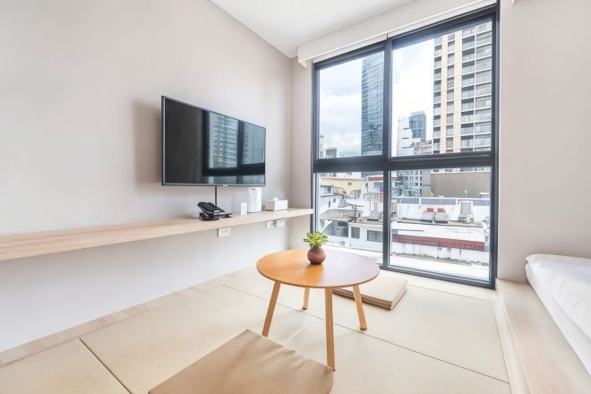 Une chambre d’hôtel moderne dotée d’une grande fenêtre, d’un téléviseur à écran plat, d’une petite table ronde agrémentée d’une plante et d’un canapé.