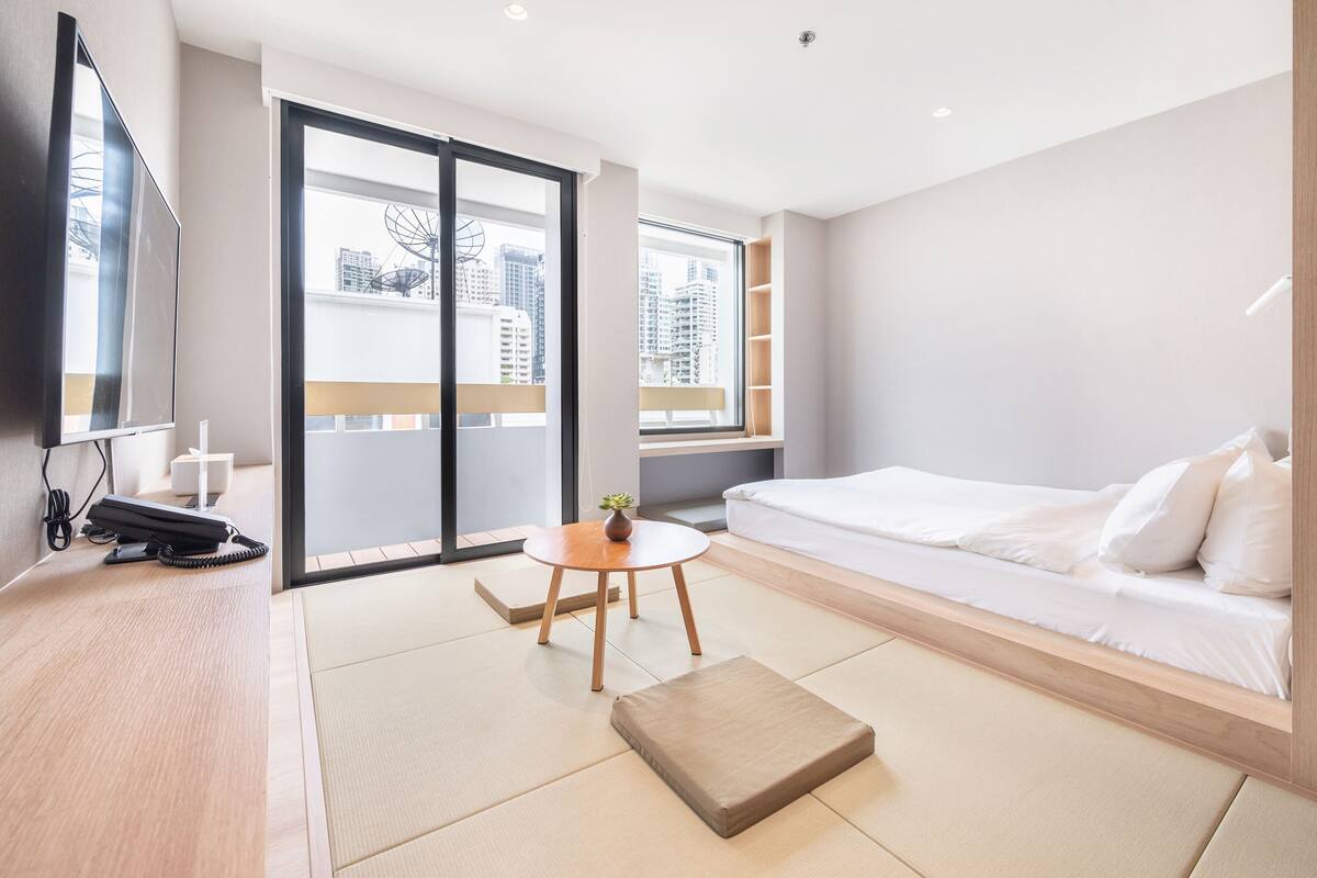A modern hotel room with a large bed, a small round table with a vase, a sofa, and a view of buildings through the window.