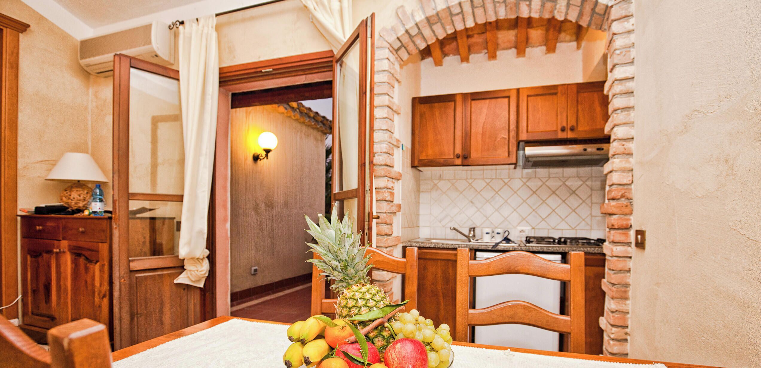 A room with a dining table set with fruits, a wooden chair, and a kitchen area with cabinets and a stove.