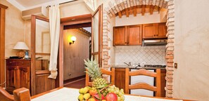 A dining area with a table set for two, featuring a fruit bowl with pineapple, grapes, and oranges. A wooden chair is visible, and the room has a kitchen in the background with wooden cabinets and a tiled backsplash.