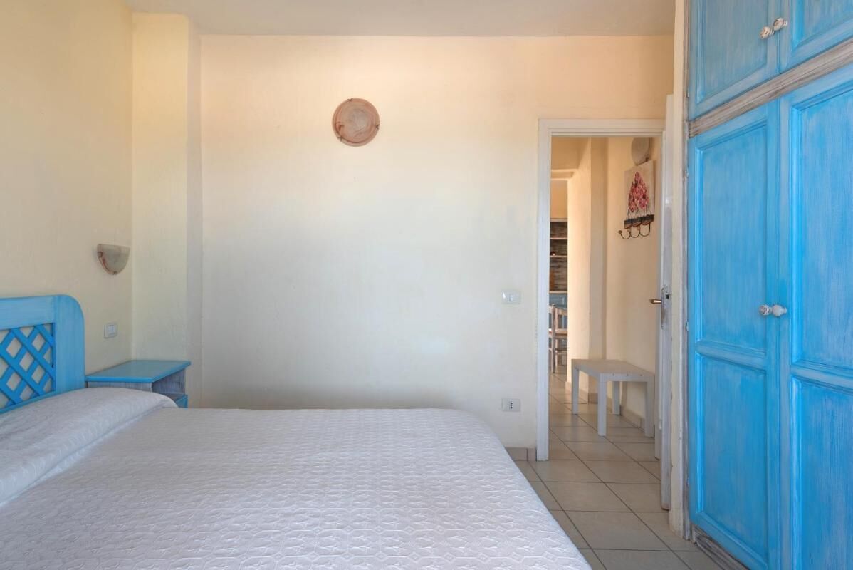Quarto com cama, mesa de cabeceira, cômoda e porta para um corredor.