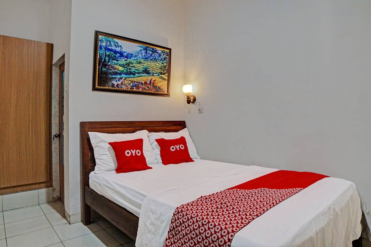 A neatly made bed with white bedding and red accents, a wooden headboard, and a wall-mounted painting above it.