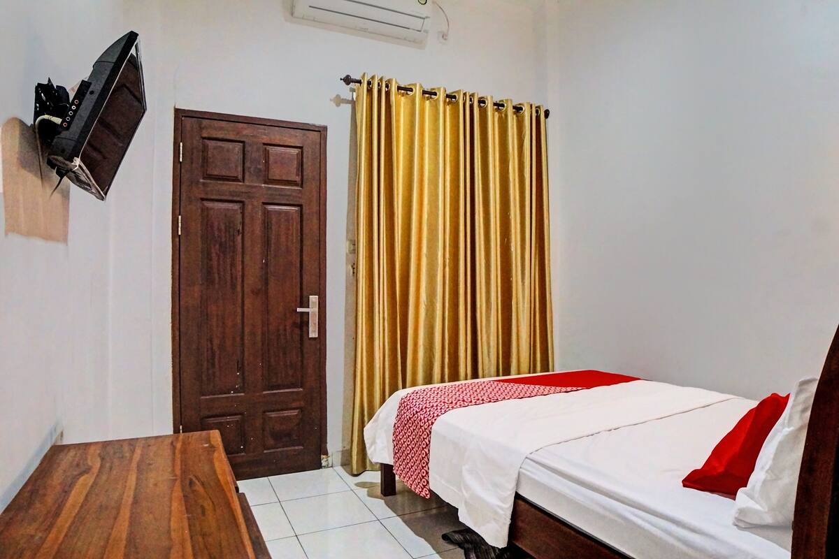 A room with a bed, a wooden table, a TV mounted on the wall, a door, and a curtain.