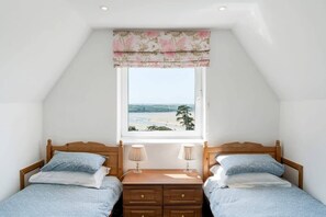 A bedroom with two beds, a window with a view of the beach, and a wooden nightstand with lamps. - Beautiful Home with Pool Overlooking Camel Estuary (Wadebridge)
