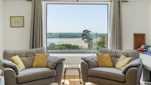 A living room with two grey sofas, a small table, and a large window with a view of the countryside. - Beautiful Home with Pool Overlooking Camel Estuary (Wadebridge)