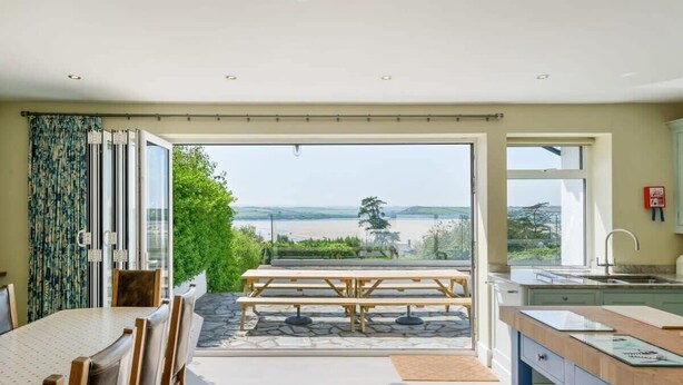A modern kitchen with a large window, a dining area with a table and chairs, and a view of a body of water. - Beautiful Home with Pool Overlooking Camel Estuary (Wadebridge)