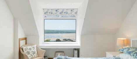 A bedroom with a large window offering a view of a beach, a bed with blue floral bedding, a chair with a floral pillow, and a bedside table with a lamp.