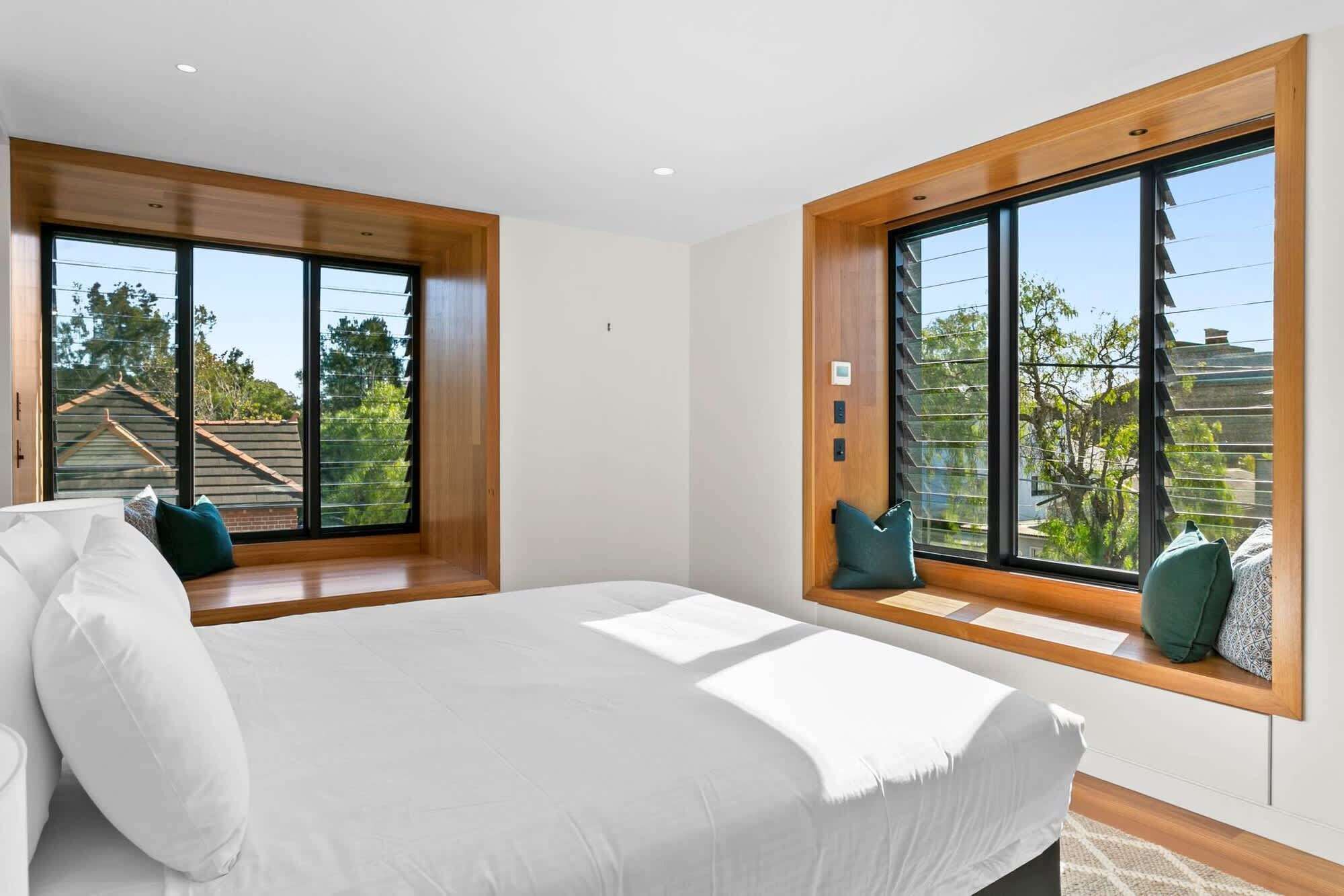 Quarto com cama grande, duas janelas com molduras de madeira e vista para o exterior.