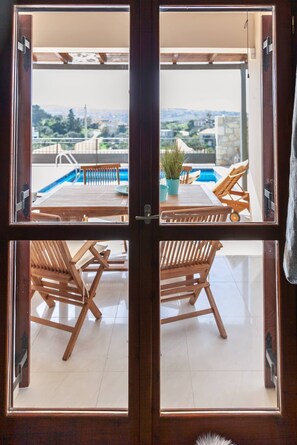 Ein Zimmer mit Holztisch und -stühlen, einer Kübelpflanze sowie Blick auf ein Schwimmbecken und die Landschaft.