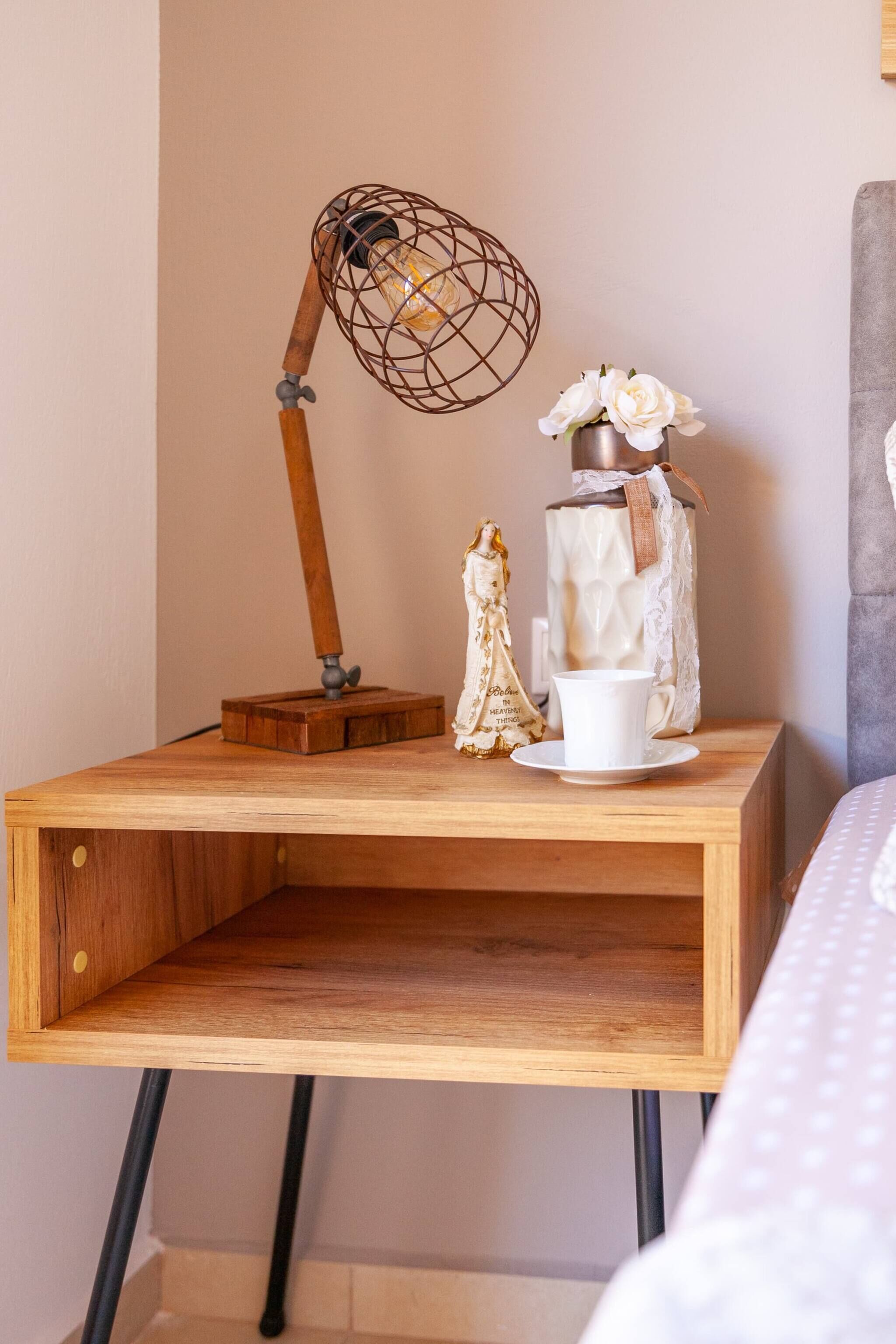 A wooden bedside table with a lamp, a vase with flowers, and a figurine.