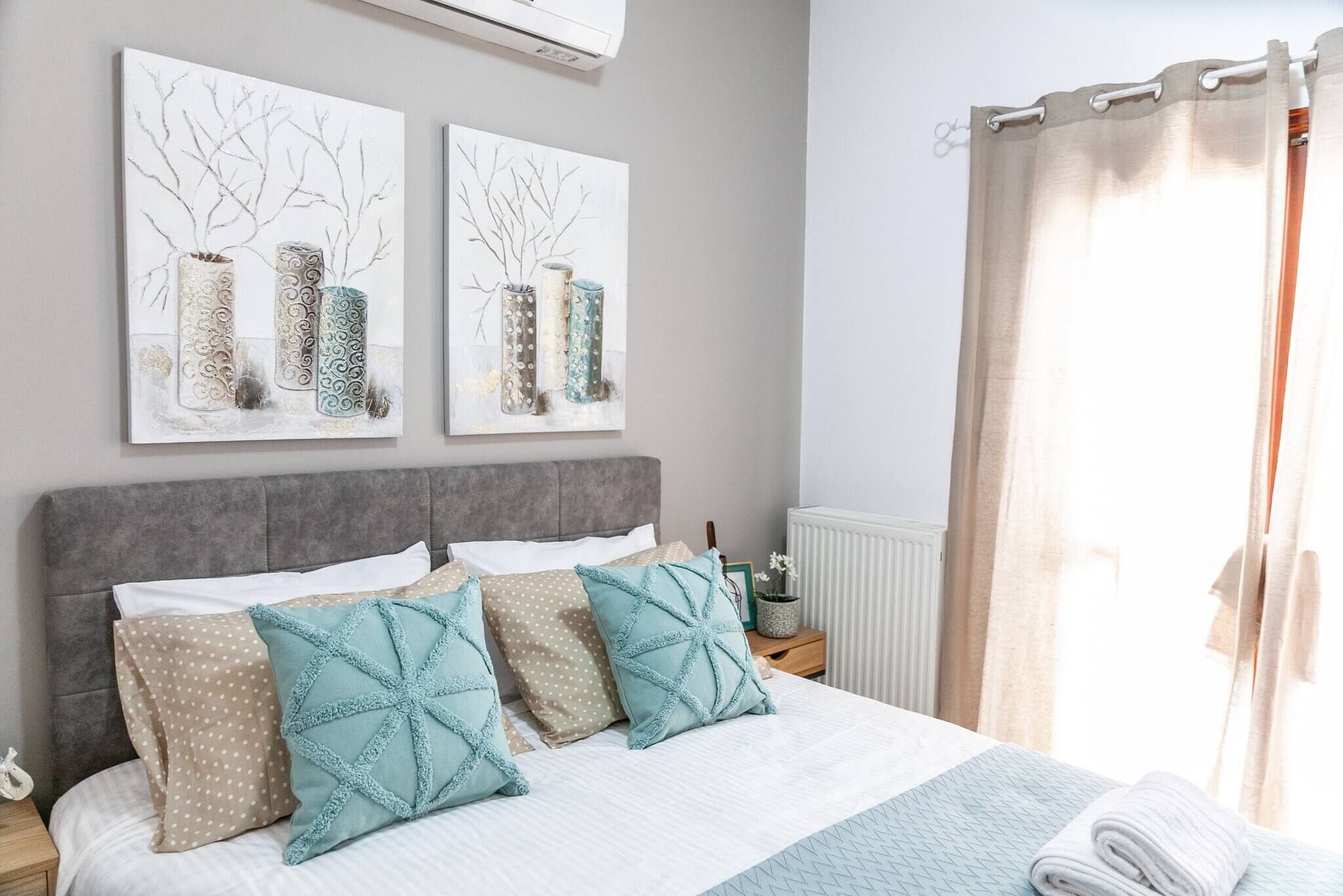 A neatly made bed with patterned pillows, a grey headboard, and two wall-mounted artworks depicting vases.