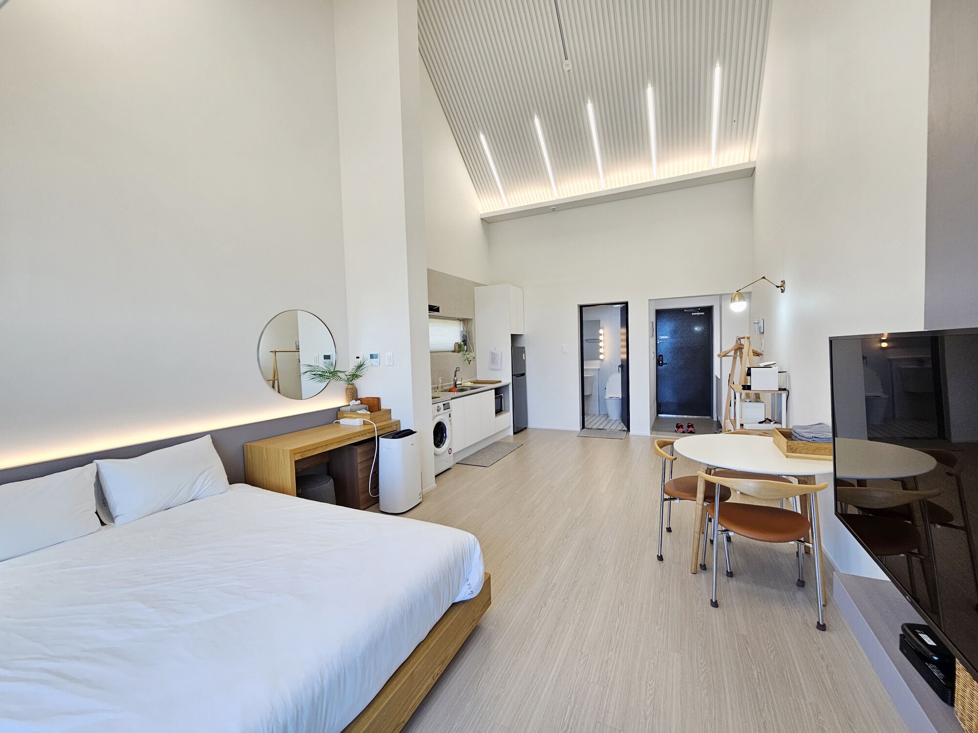 A modern hotel room with a bed, a desk with a mirror, a dining area with chairs, a TV, and a kitchenette.