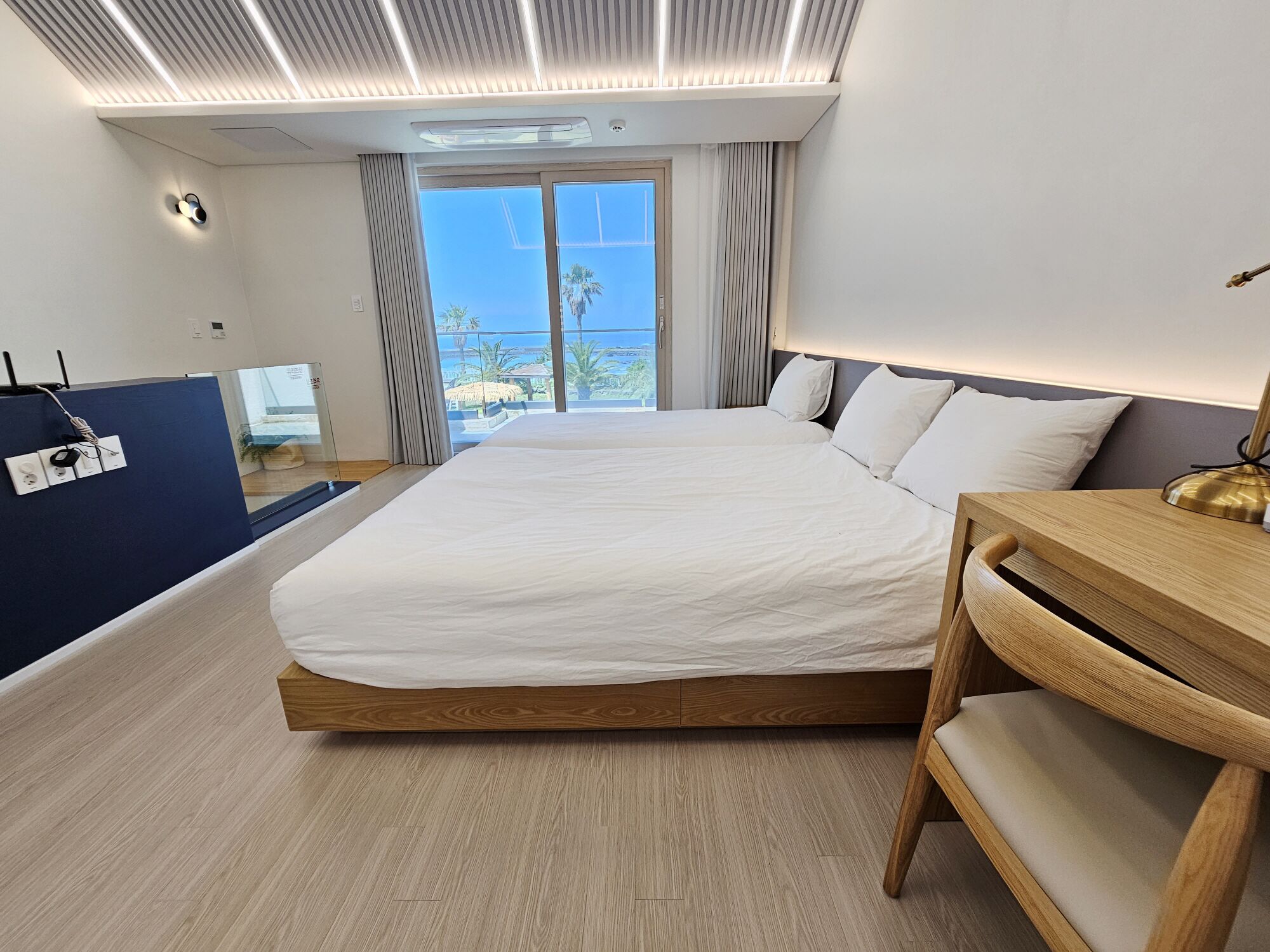 Une chambre d’hôtel moderne avec un grand lit, une tête de lit en bois, une table de chevet et une vue sur l’océan.