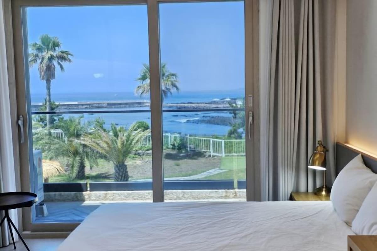 A hotel room with a large sliding glass door leading to a balcony with a view of the ocean and palm trees.