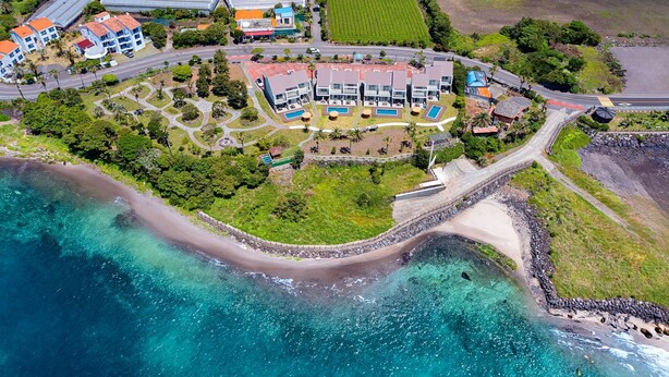 Vista aérea de un complejo turístico frente al mar, con varios edificios y un jardín bien cuidado.