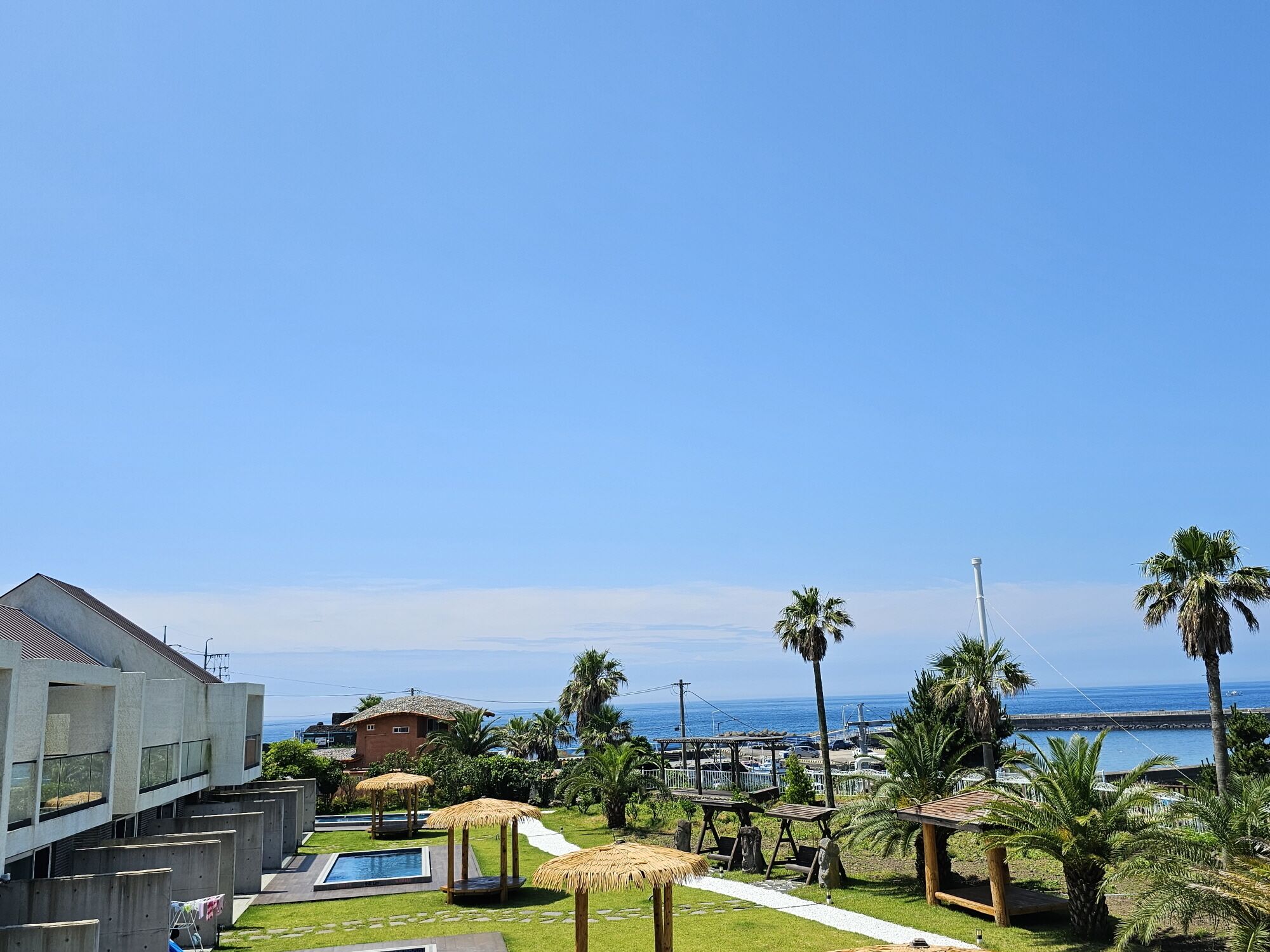 A resort with a swimming pool, thatched-roof structures, palm trees, and a view of the ocean.