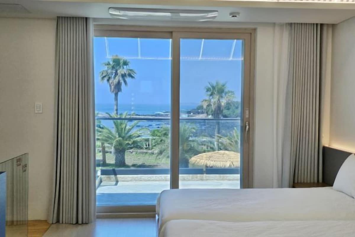 A hotel room with a bed, a large window with curtains, and a view of palm trees and the ocean.