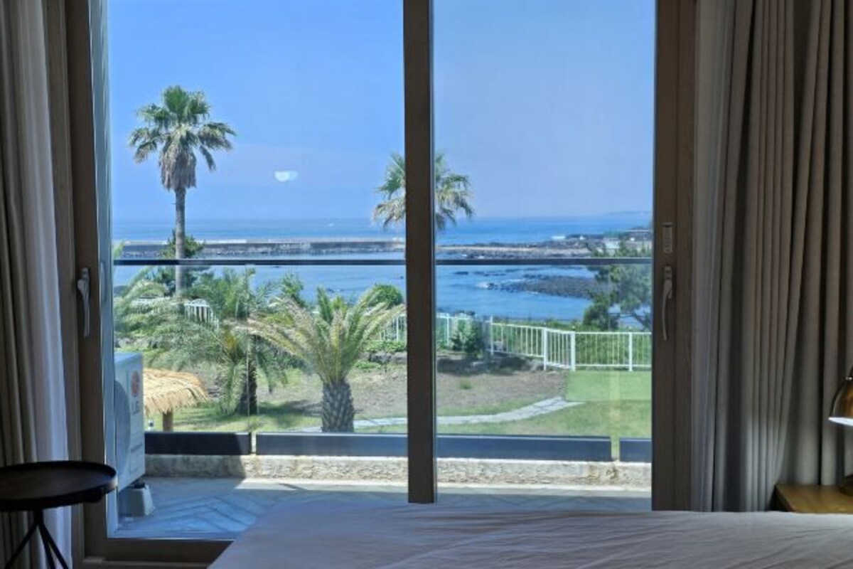 A room with a large window overlooking a beach, palm trees, and a clear sky.