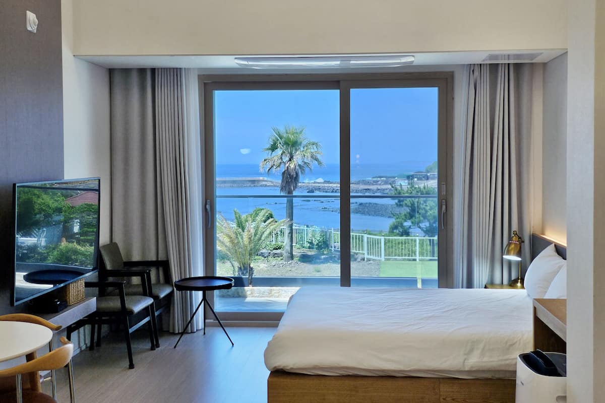 A hotel room with a large bed, a TV, a desk with a chair, and a balcony with a view of the sea and palm trees.
