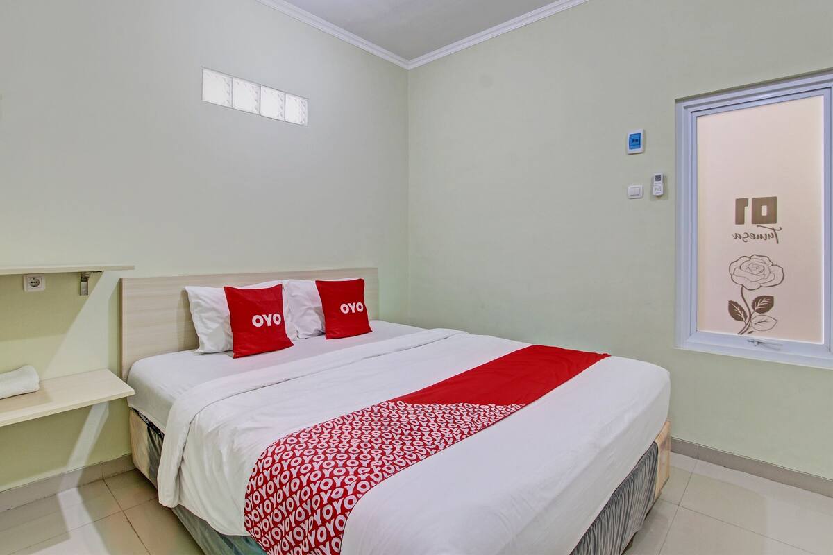 A neatly made bed with red and white bedding, a bedside table with a white towel, and a window with a floral design.