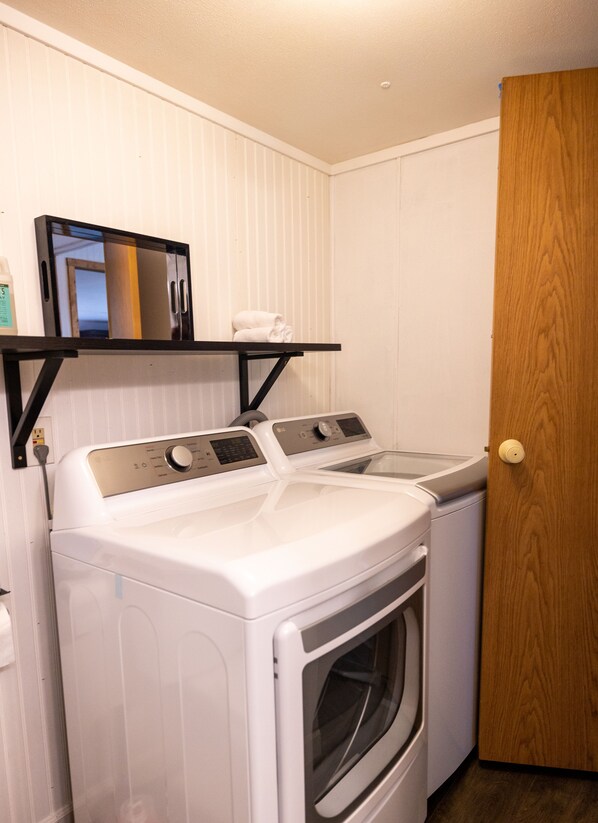 A laundry area with a front-loading washer and dryer, a shelf with a mirror and a bottle, and a wooden door.