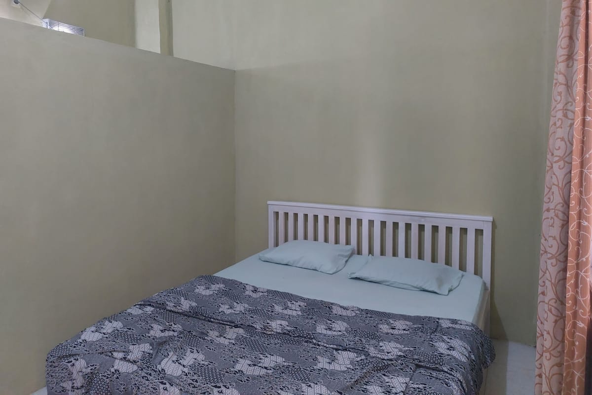 A small, single-bed room with a white headboard, a patterned bedspread, and a plain wall.