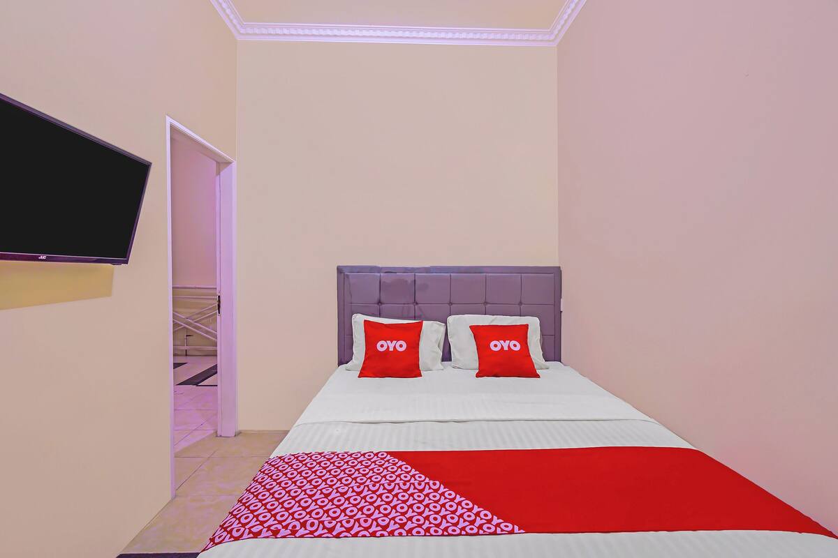 Een slaapkamer met een bed, twee rode kussens met de tekst 'OYO', een aan de muur bevestigde televisie en een deur die leidt naar een trap.