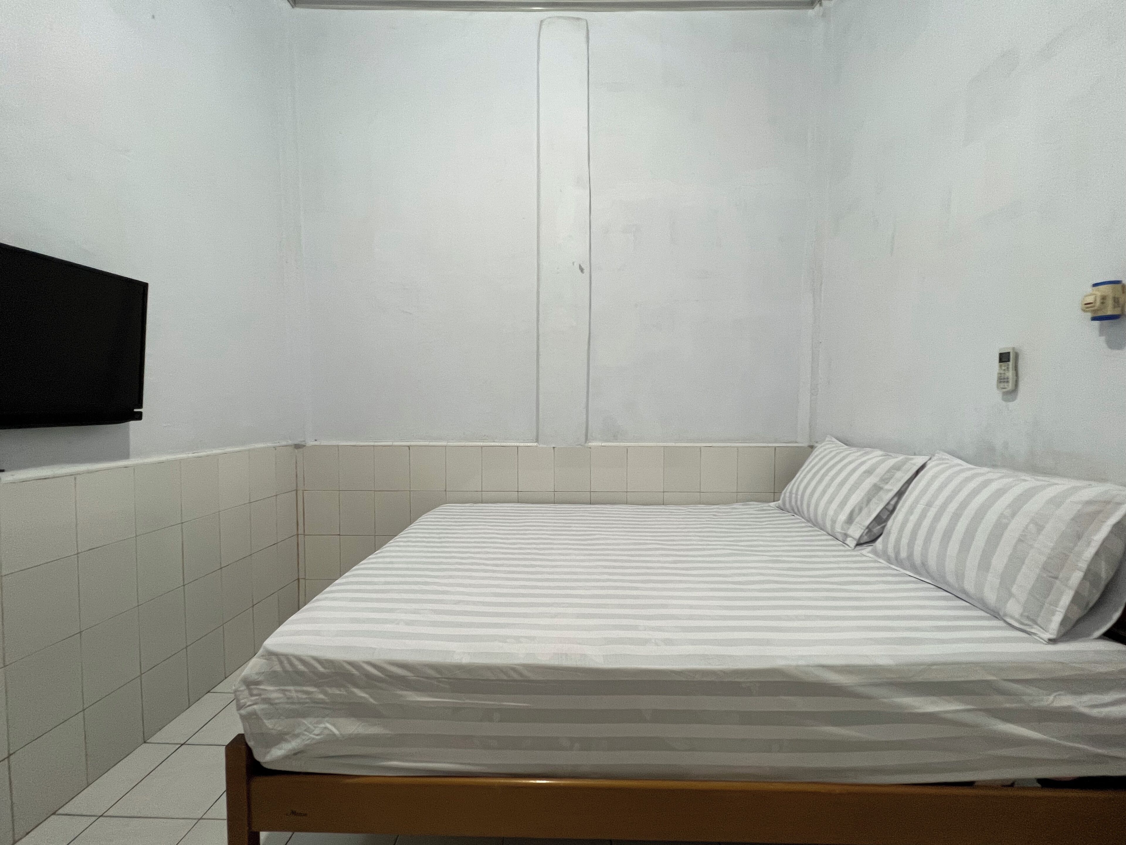 A small, single-bed room with white walls and a tiled floor. There is a television mounted on the wall to the left of the bed.