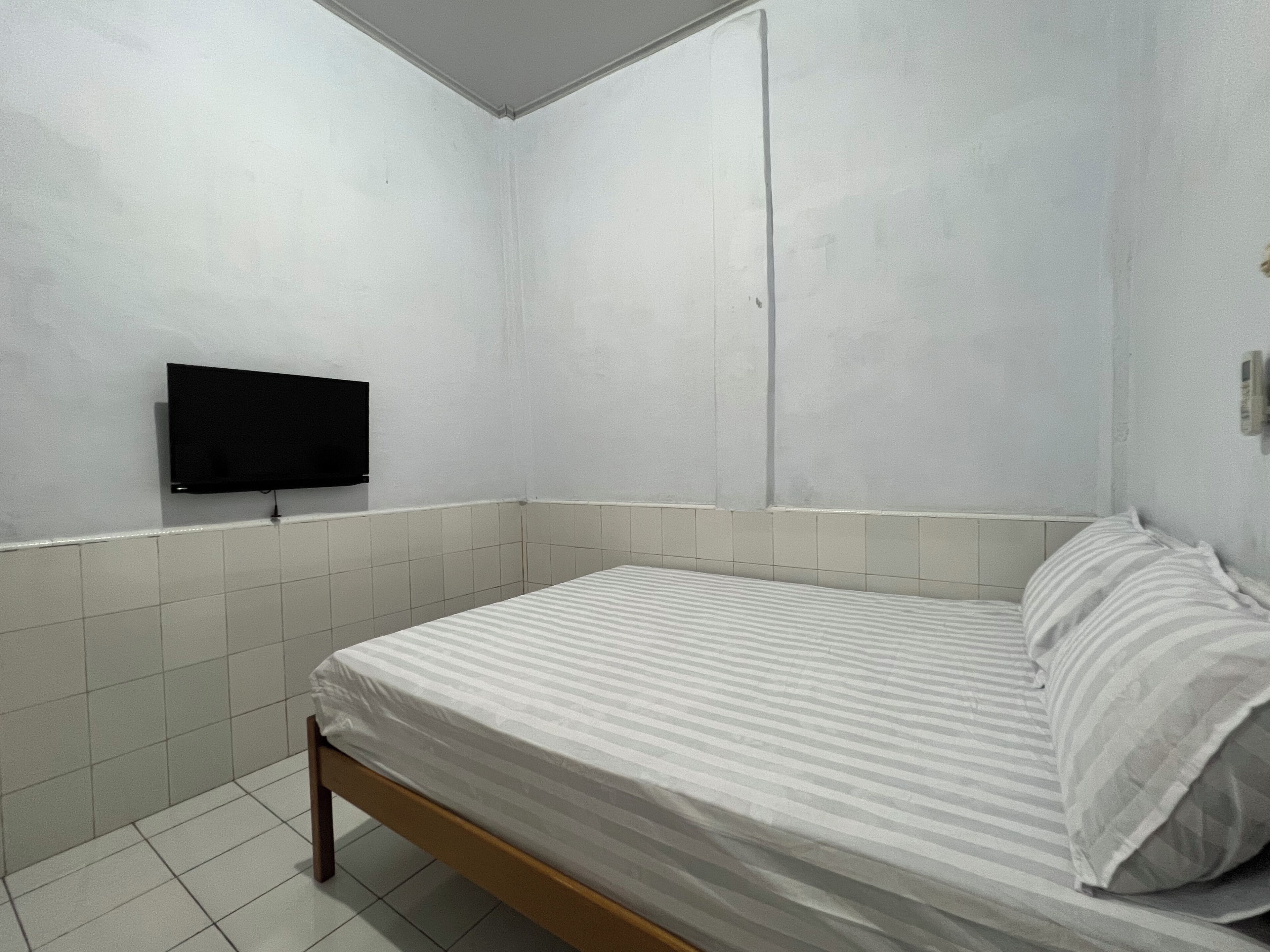 A small, clean room with a single bed, a flat-screen TV mounted on the wall, and a tiled floor.
