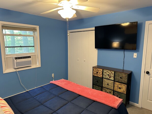 A bedroom with a bed, a ceiling fan, a window with blinds, a TV on the wall, and a dresser.