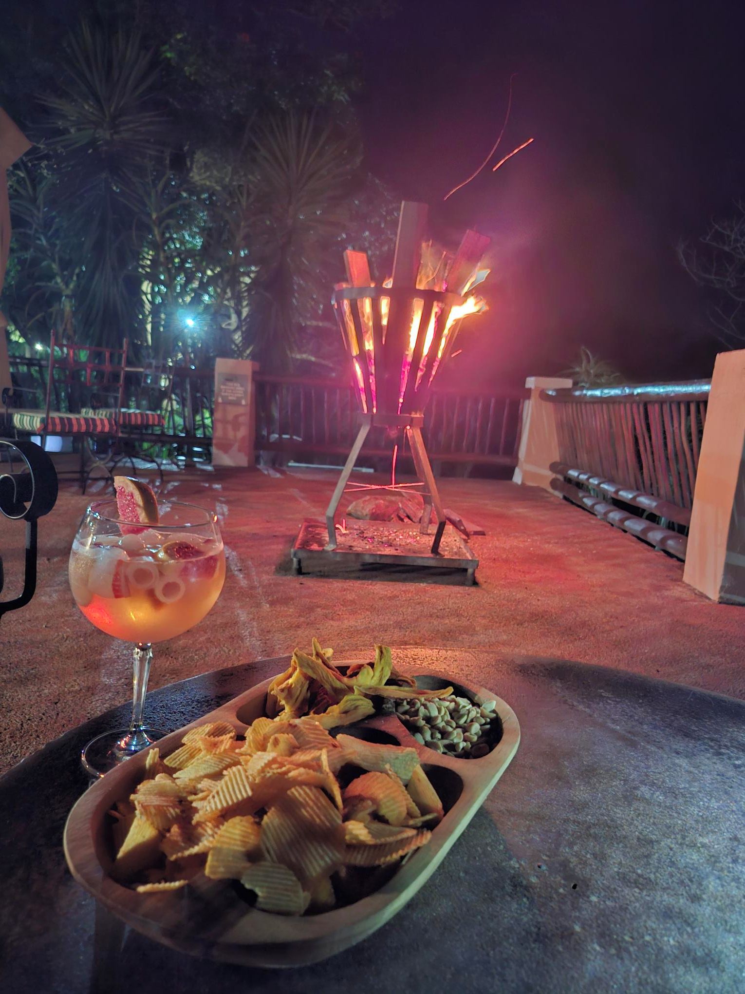 A table with a drink and snacks, a lit fire feature, and a view of trees at night.