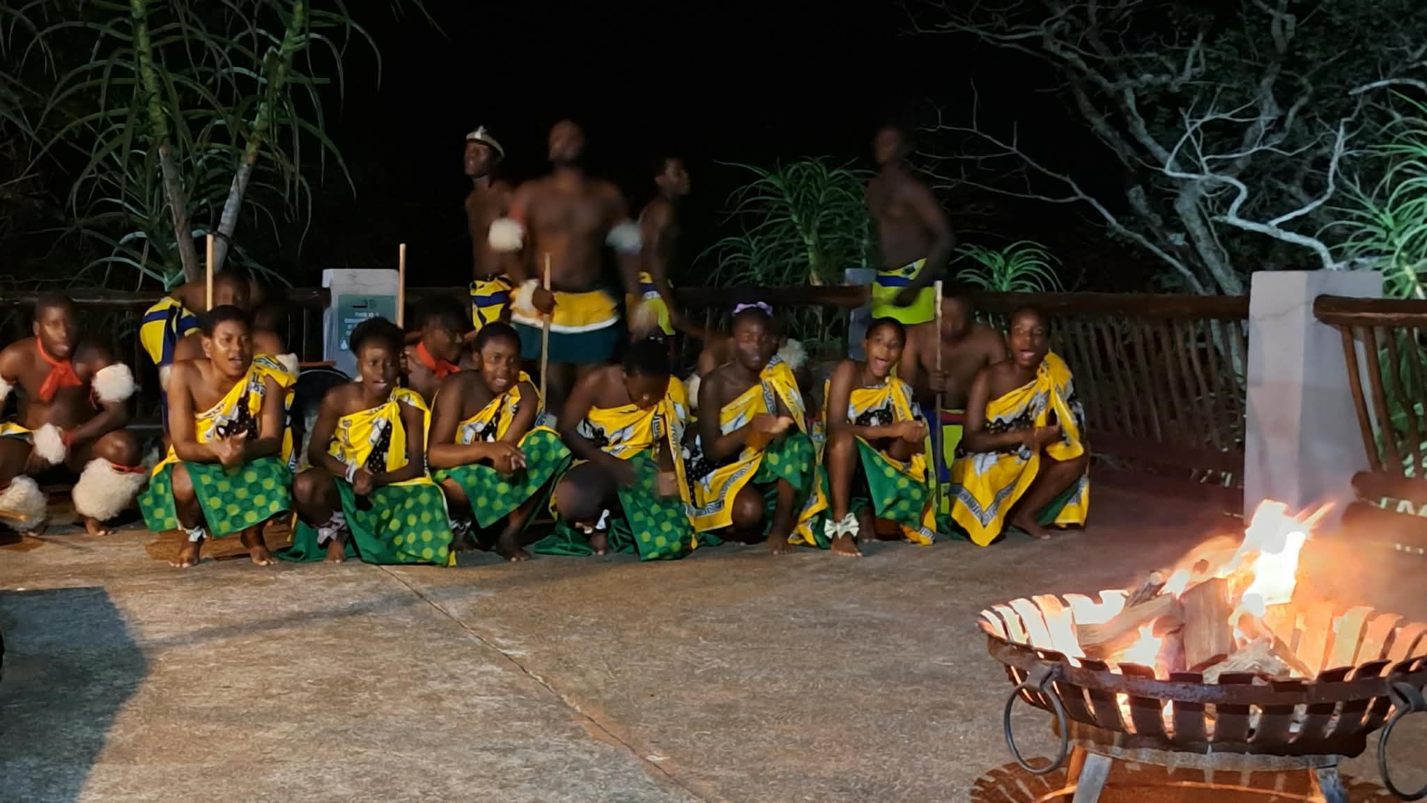 A group of people seated outdoors at night, some wearing traditional attire, with a fire in the foreground.