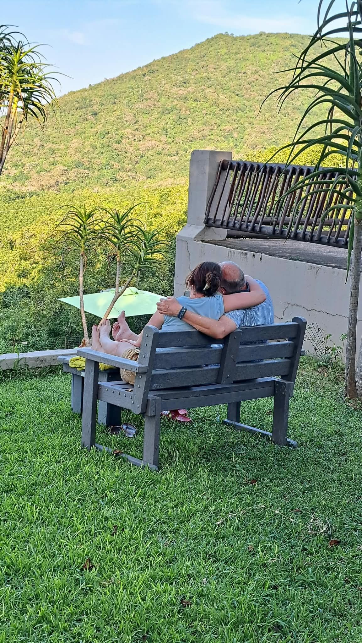 Ein Paar sitzt auf einer Bank in einer grünen Parklandschaft, im Hintergrund ist ein Hügel zu sehen.