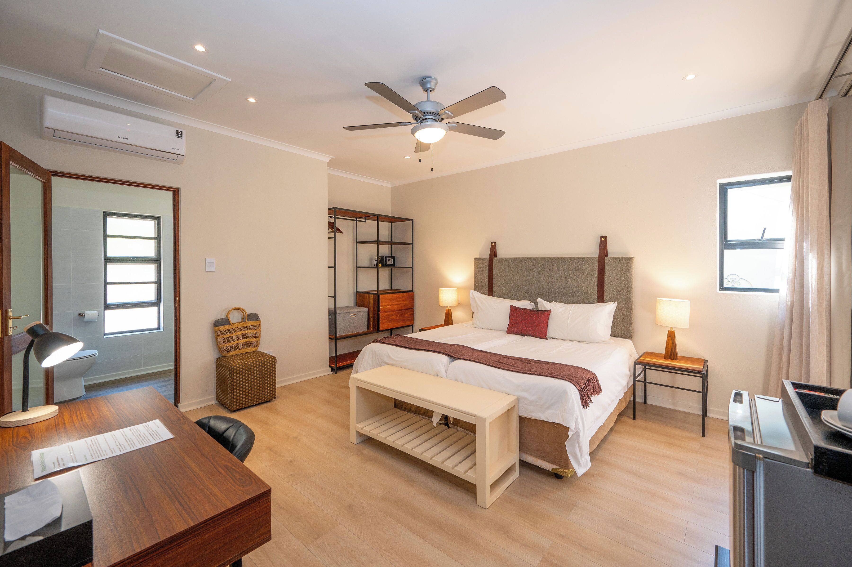 A hotel room with a bed, bedside tables, a desk, a chair, a ceiling fan, an air conditioner, a window, and a bathroom visible through an open door.