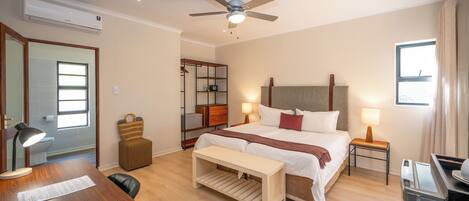 A hotel room with a bed, bedside tables, a desk, a chair, a ceiling fan, an air conditioner, a window, and a bathroom visible through an open door.