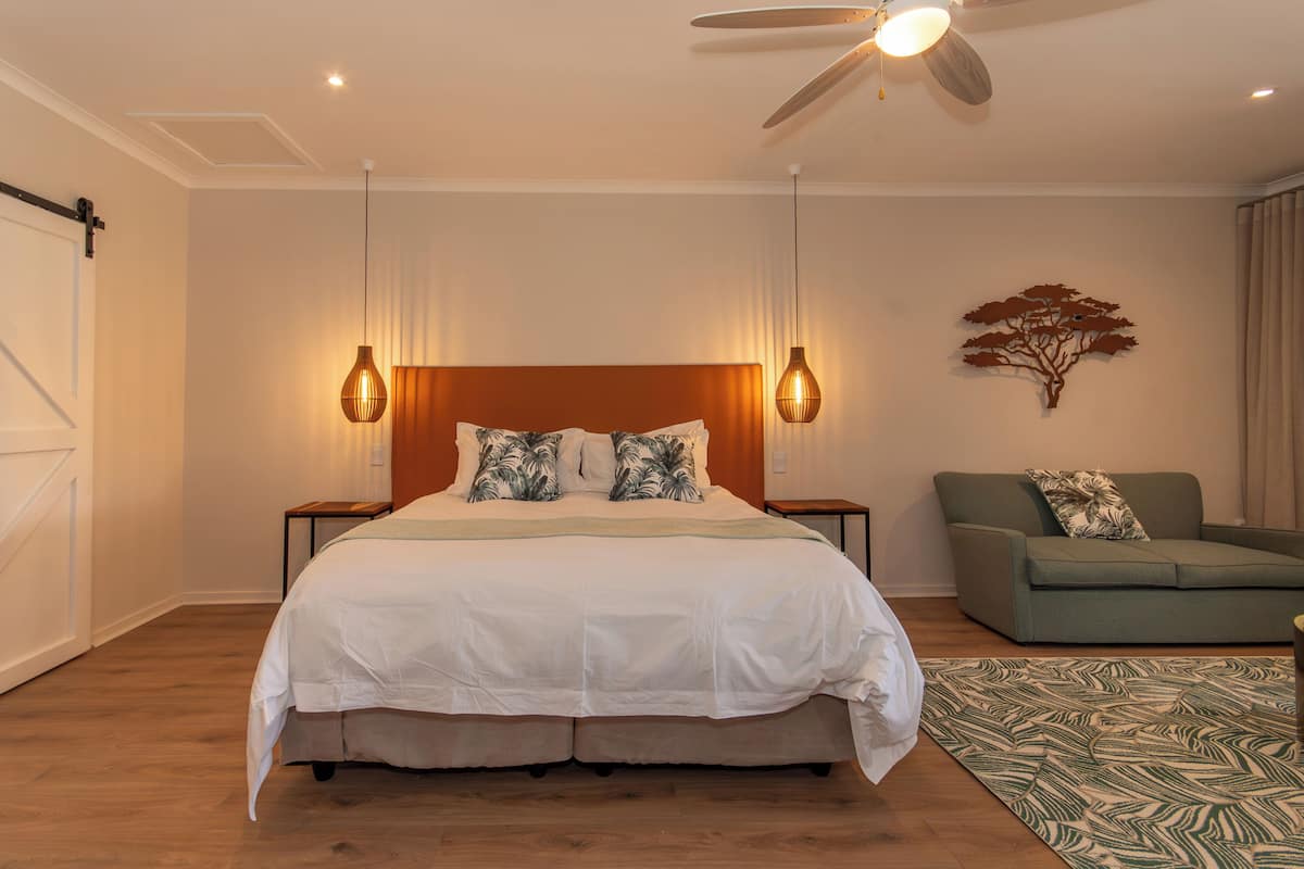 A bedroom with a large bed, a ceiling fan, two bedside tables, a green sofa, and a patterned rug.