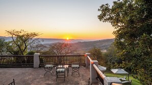 Une terrasse avec des meubles d’extérieur, une vue sur le coucher de soleil au-dessus d’une vallée et une balustrade en bois.