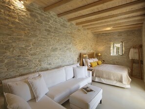 A bedroom with a stone wall, wooden ceiling beams, a white bed, a white sofa, and a small table with a book and glass.