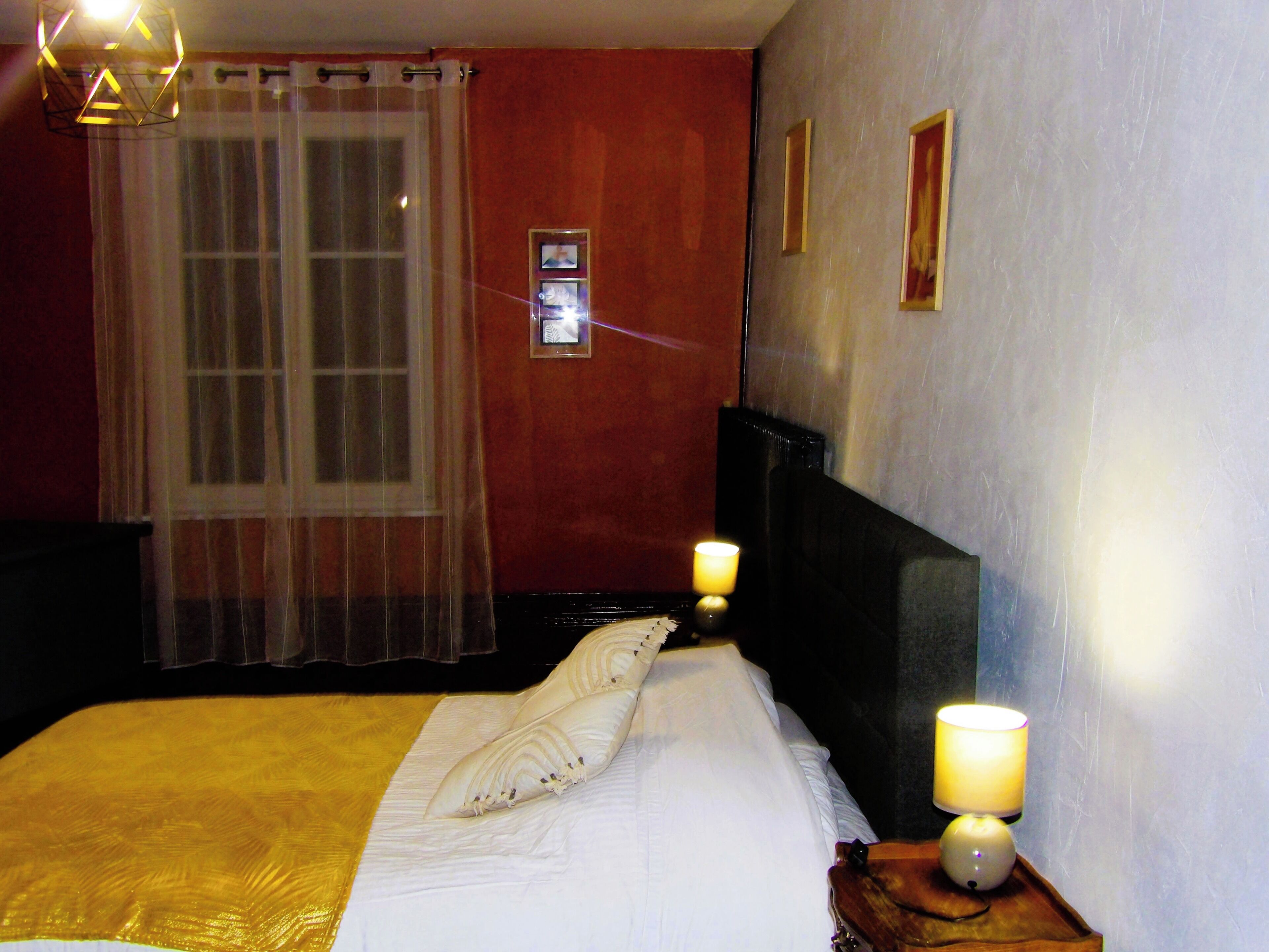 A hotel room with a bed, two bedside lamps, a window with sheer curtains, and a wall with framed pictures.
