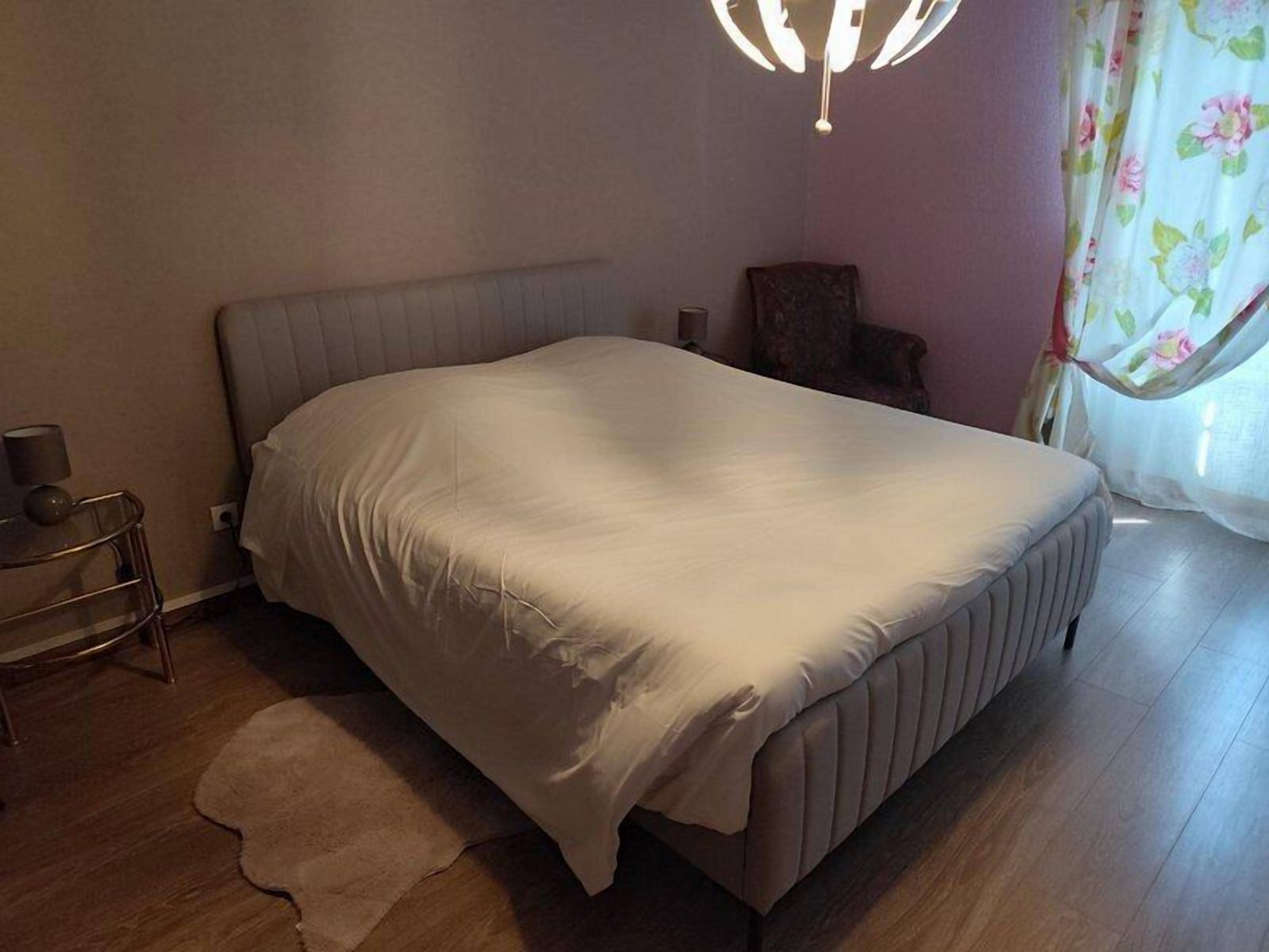 A bedroom with a bed, a nightstand, a chair, a lamp, and a window with curtains.