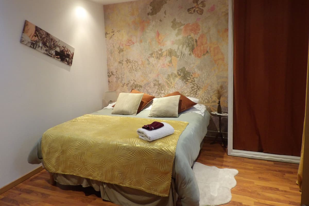 Une chambre à coucher avec un lit, des oreillers, une table de chevet, une lampe, un tableau au mur et un rideau.